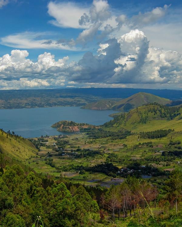The green caldera rim of Lake Toba dropping to the water in North Sumatra, with a cultivated valley floor below and cumulus cloud over the far shore