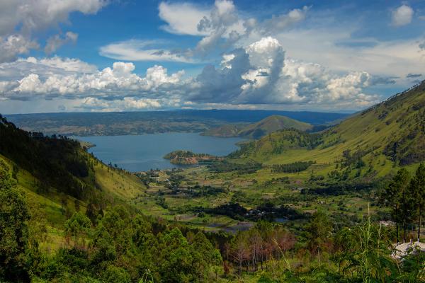 The green caldera rim of Lake Toba dropping to the water in North Sumatra, with a cultivated valley floor below and cumulus cloud over the far shore