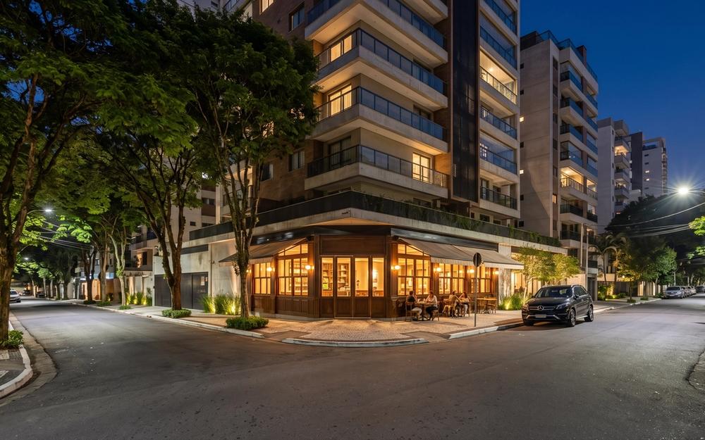 Esquina residencial em Moema ao anoitecer: prédio contemporâneo com varandas iluminadas, café de esquina em madeira, árvores na calçada