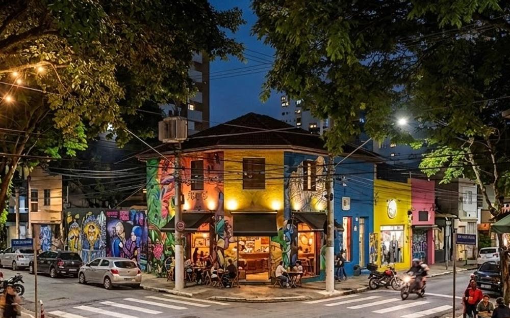 Esquina da Vila Madalena à noite: sobrado coberto de grafites, bares com mesas na calçada e luzes de festa entre as árvores