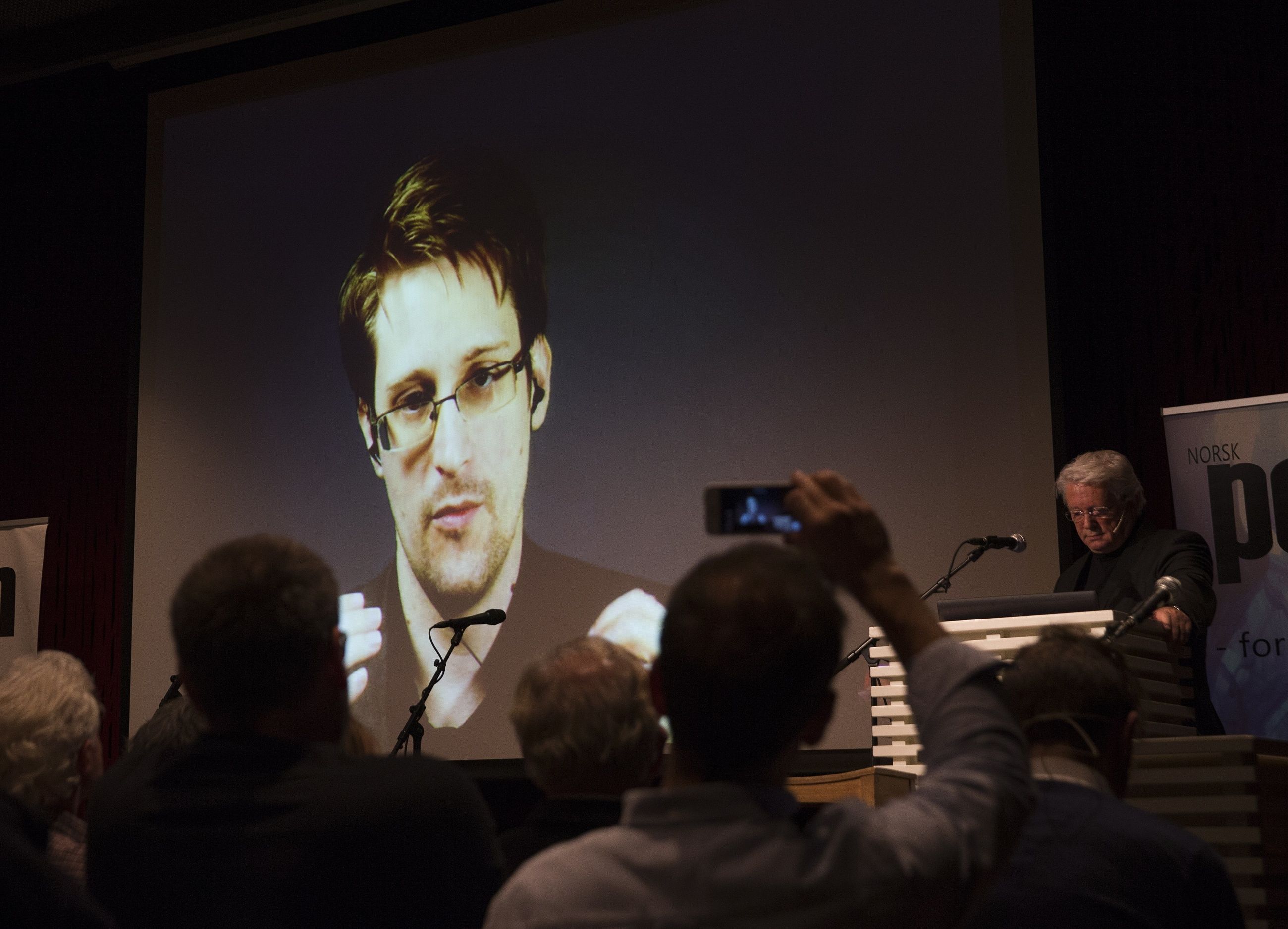 Edward Snowdens ansikt vises på en storskjerm med i en sal med mennesker som ser på og tar bilder med mobiltelefon i forgrunnen. (fra et arrangement på Litteraturhuset i Oslo i 2016. FOTO: Berit Roald, NTB)