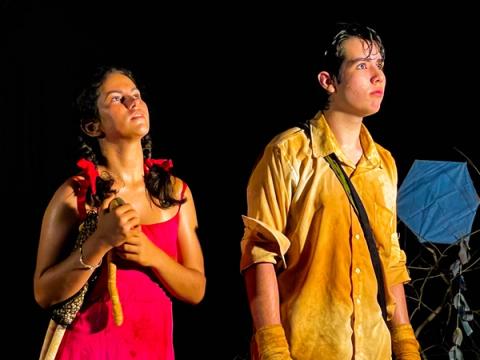 Atriz com vestido vermelho segurando boneca, ao lado de ator em cenário escuro com uma pipa ao fundo.