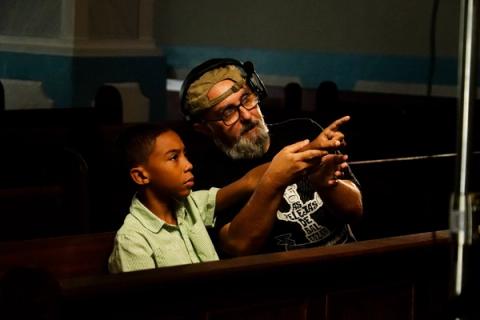 O diretor, usando fones e óculos, orienta um jovem ator sentado ao seu lado em um banco de madeira, apontando para fora da cena.