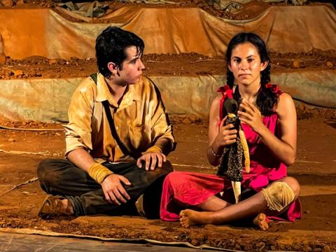 Ator e atriz sentados no chão conversando, ela segura uma boneca.