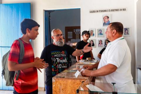 Diretor conversa com jovem ator em balcão de museu. Fotos de diretores na parede ao fundo.