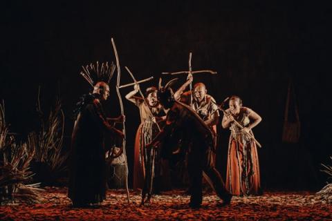 Grupo de atores em cena teatral, vestidos com trajes tradicionais e segurando varas em formas de cruz.