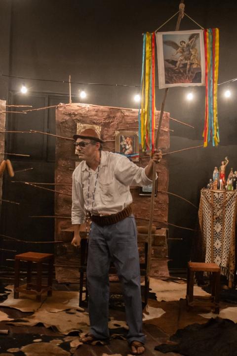 Ator segurando uma haste com imagem religiosa pendurada, em cenário teatral.