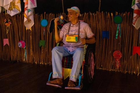 Ator sorridente, vestindo chapéu e bolsa colorida, sentado em cadeira de rodas decorada.