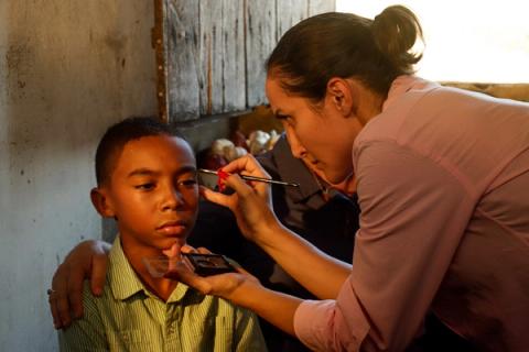 Uma maquiadora aplica maquiagem com um pincel no rosto de um menino sentado, que olha para o lado, em preparação para a gravação de uma cena.
