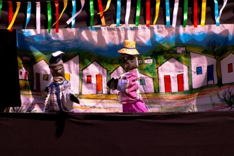 Bonecos de teatro interagindo em cena com fundo de casas rurais.
