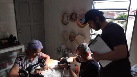 Mulher costurando enquanto dois homens observam e fotografam.