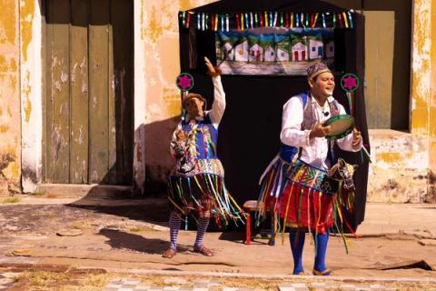 Dois artistas vestidos coloridamente ao lado de um palco de bonecos.