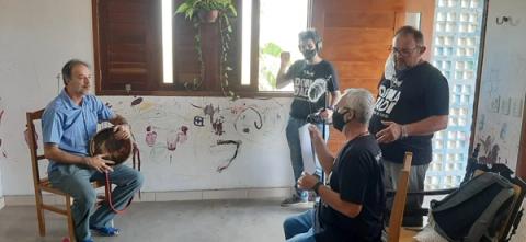 Homem tocando tambor enquanto concede entrevista fala para grupo de filmagem.
