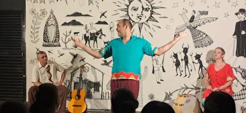 Três artistas com instrumentos em um palco decorado com motivos culturais diante do público.