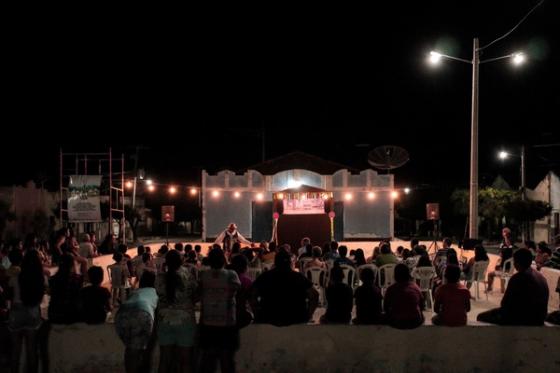 Apresentação ao ar livre à noite, com plateia em cadeiras plásticas e palco iluminado por luzes. Um artista se destaca no centro do palco.