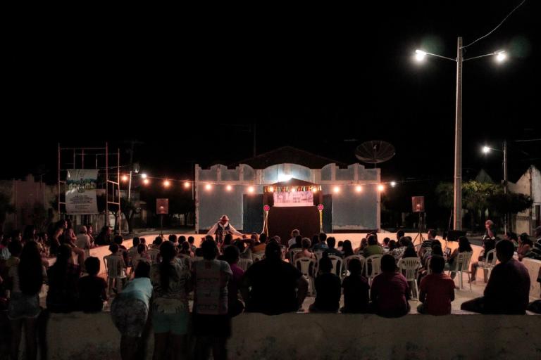 Apresentação ao ar livre à noite, com plateia em cadeiras plásticas e palco iluminado por luzes. Um artista se destaca no centro do palco.