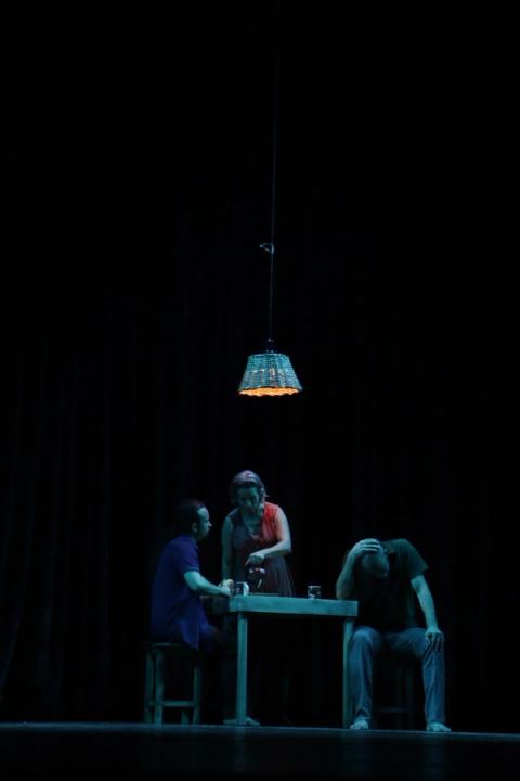 Três pessoas em torno de uma mesa, iluminadas por uma lâmpada pendente em palco escuro.