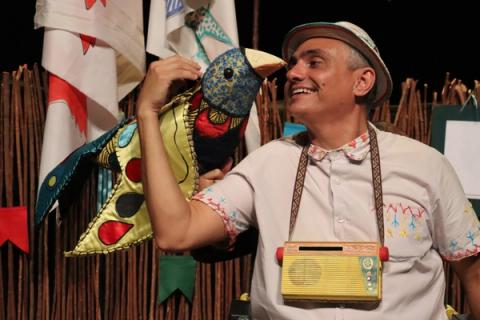 Ator segurando marionete de pássaro, sorrindo em cenário rústico.