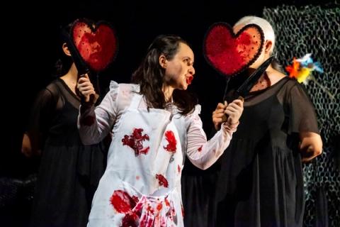 Atriz central com vestido branco manchado de sangue em cena dramática com dois atores laterais segurando corações vermelhos e facas.