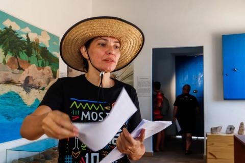 Produtora segura papéis em salão de museu com painel de paisagem marinha ao fundo.