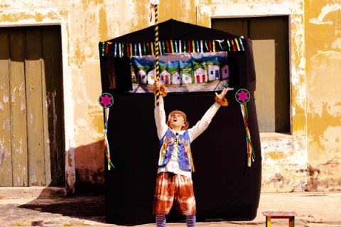 Ator vestindo traje colorido, em frente a um palco de marionetes ao ar livre.