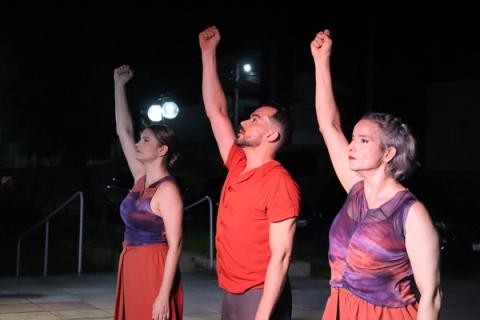 Grupo de três bailarinos levantando os braços no ar, dançando ao ar livre à noite.