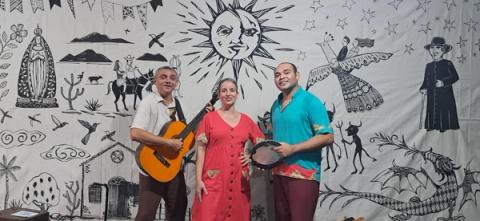 Três artistas com instrumentos em um palco decorado com motivos culturais diante do público.