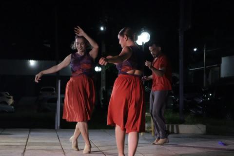 Grupo de bailarinos dançando ao ar livre à noite, com luzes de rua ao fundo.