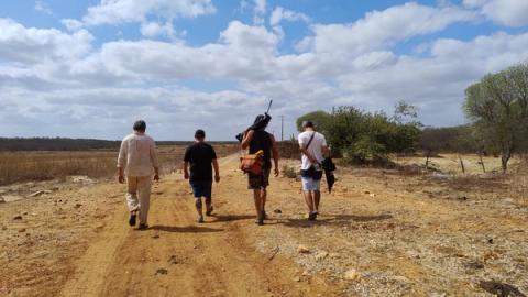 Quatro pessoas caminhando por estrada rural, carregando equipamentos.