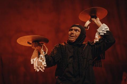 Personagem teatral com adereços, segurando objetos circulares em fundo vermelho.