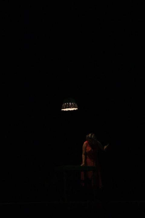 Bailarina em vestido vermelho, em cena com lâmpada pendente e fundo escuro.