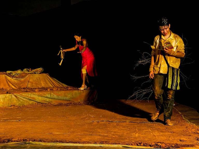 Atriz em vestido vermelho segura boneca, enquanto ator lê livro.