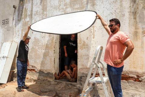 Dois membros da equipe usam um grande rebatedor para controlar a luz sobre um menino sentado na soleira de uma porta, durante as filmagens.