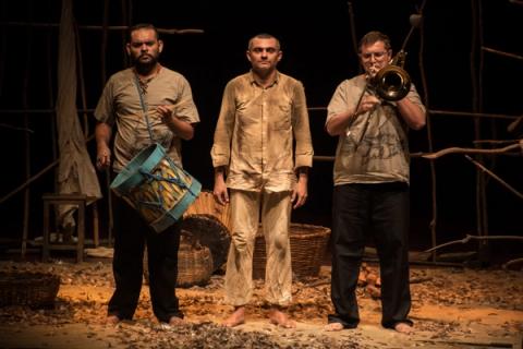 Três atores em cena, um tocando tambor, outro segurando trombone e o terceiro imóvel no centro.