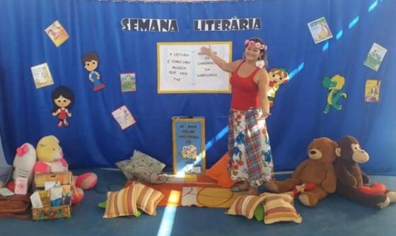 Mulher em frente a painel com tema "Semana Literária", rodeada de almofadas e brinquedos.