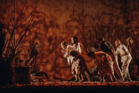 Grupo de atores em cena teatral, vestidos com roupas simples e carregando varas, sob iluminação vermelha.