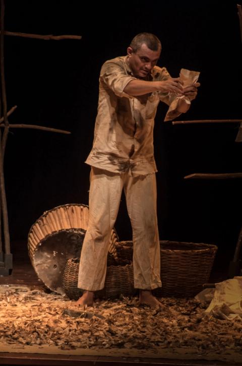 Ator examinando objeto com atenção, cenário rústico ao fundo.