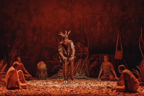 Cena teatral com personagem central usando chifres, rodeado por outros atores sentados no chão.