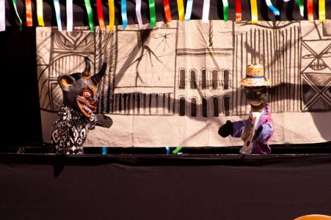 Dois bonecos de teatro em cena com fundo abstrato e detalhes geométricos.