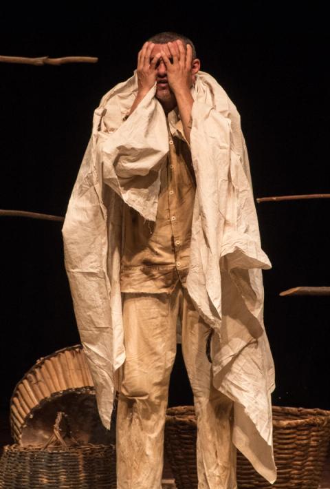 Ator cobrindo rosto com mãos, vestido com pano branco, em cenário sombrio.