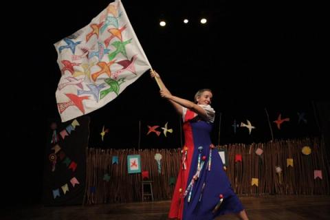 Atriz segurando bandeira com desenhos de pássaros coloridos, dançando em palco decorado.