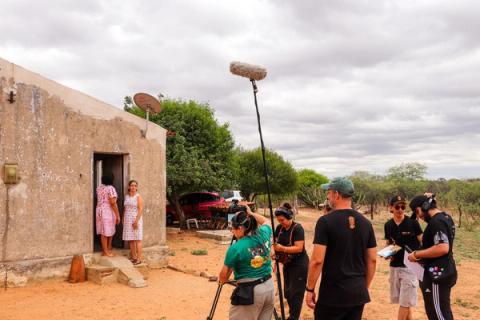 Equipe de filmagem em uma locação externa rural, com o operador de boom, diretor e outros membros se preparando para gravar com duas atrizes na porta de uma casa.