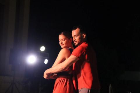 Casal de bailarinos dançando juntos em palco escuro, iluminados por holofotes.