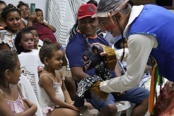 Um artista vestido com traje colorido e máscara interage com uma criança sentada em cadeira plástica, enquanto outras crianças e adultos observam atentamente.