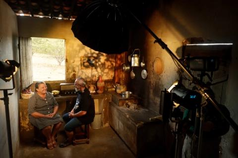 Em um set montado como uma cozinha antiga, o diretor conversa com uma atriz sentada, com equipamentos de iluminação e câmera ao redor.