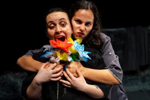 Duas atrizes em cena teatral: uma à frente com expressão de surpresa segura buquê de flores coloridas, enquanto outra a abraça por trás com expressão intensa.