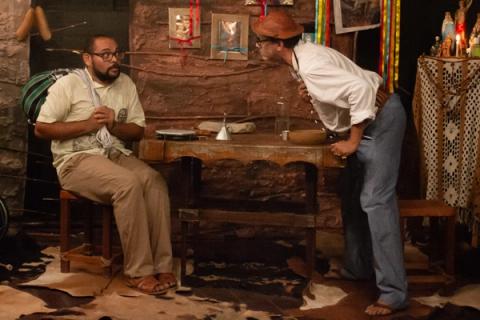 Dois atores interagindo em cena teatral ao redor de uma mesa.