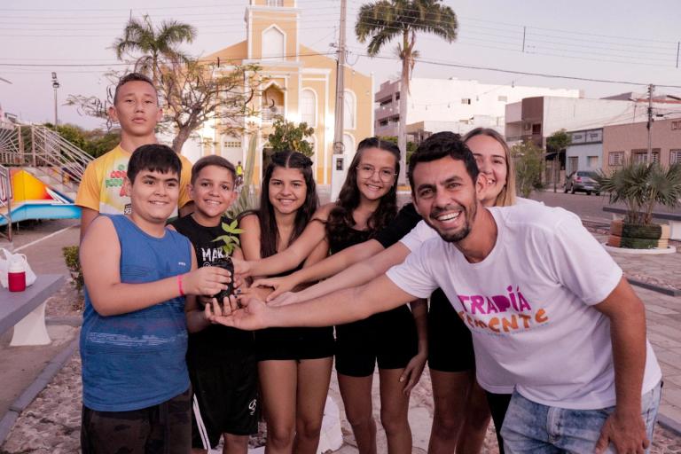 Pessoas segurando uma muda de planta juntas, simbolizando o crescimento da semente e união.