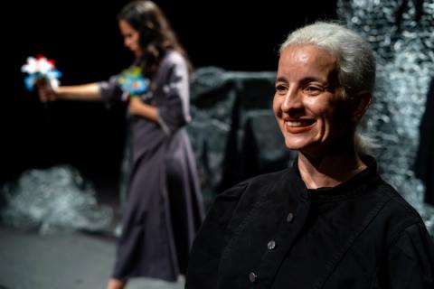 Uma mulher de cabelos brancos e roupa preta sorri para a câmera. Ao fundo, desfocada, outra mulher de vestido cinza se afasta segurando um buquê de flores coloridas.