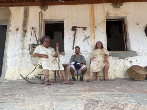 Três pessoas sentadas em varanda rústica com ferramentas na parede.